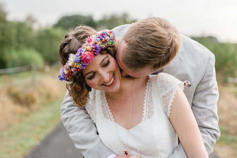 Die Braut lächelt bei den Küssen ihres Bräutigams und trägt einen Blumenkranz - Fotografin: Sonja Yasmin Fotografie, Hochzeitsfotografin in Köln, Bonn und ganz NRW, Fotografin für Portraitfotos und Familienfotos