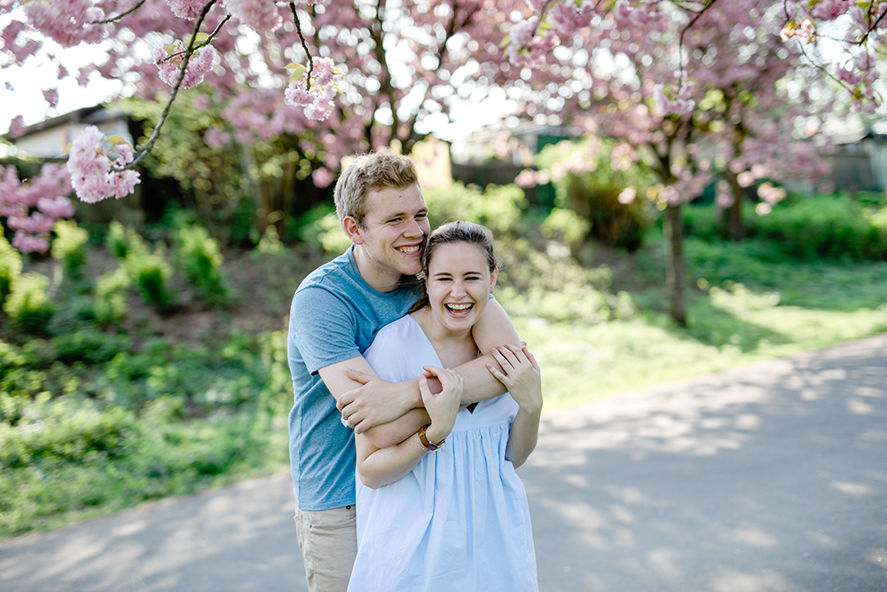 Das Paar lacht auf dem Weg umgeben von Kirschblüten - Fotografin: Sonja Yasmin Fotografie, Hochzeitsfotografin in Köln, Bonn und ganz NRW, Fotografin für Portraitfotos und Familienfotos