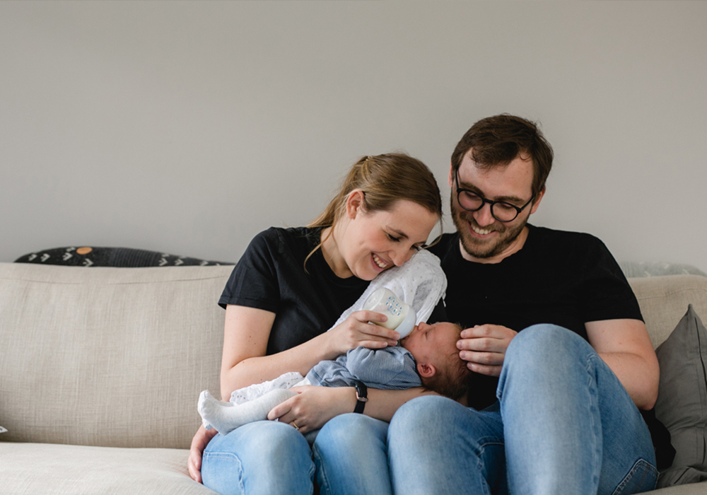 Die Eltern füttern ihr Baby auf der Couch während des Familienshootings zu Hause - Fotografin: Sonja Yasmin Fotografie, Hochzeitsfotografin in Köln, Bonn und ganz NRW, Fotografin für Portraitfotos und Familienfotos