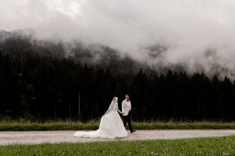 Das Brautpaar schlendert entspannt einen Weg in den Bergen von Südtirol entlang. Im Hintergrund vor den dunklen Bäumen Bilder sich Nebel - Fotografin: Sonja Yasmin Fotografie, Hochzeitsfotografin in Köln, Bonn und ganz NRW"